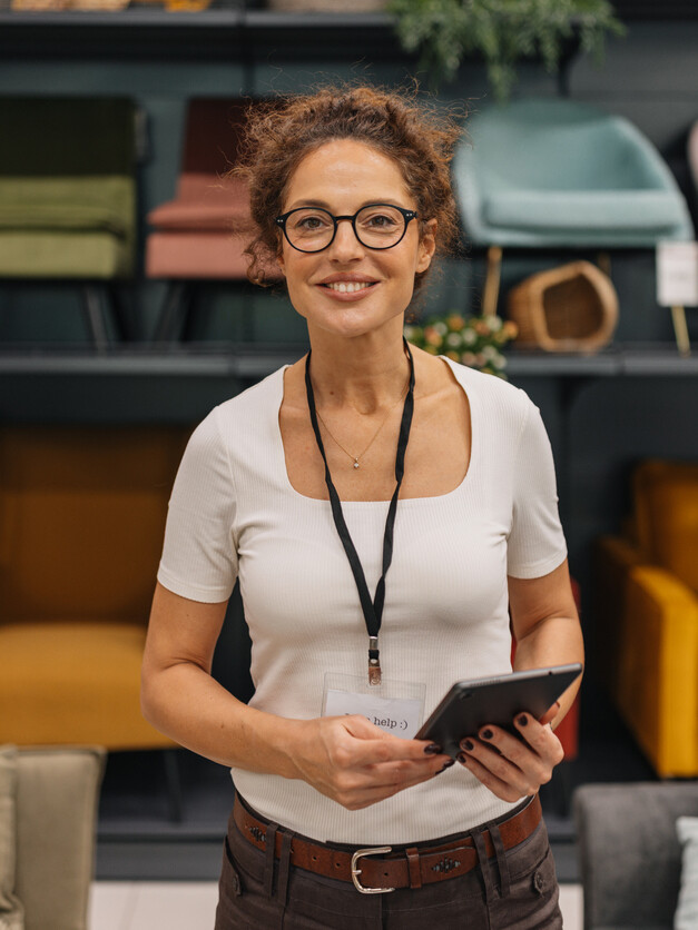 A smiling woman stands in a modern furniture store, holding a tablet while surrounded by colorful seating options, demonstrating her enthusiasm and engagement in retail.