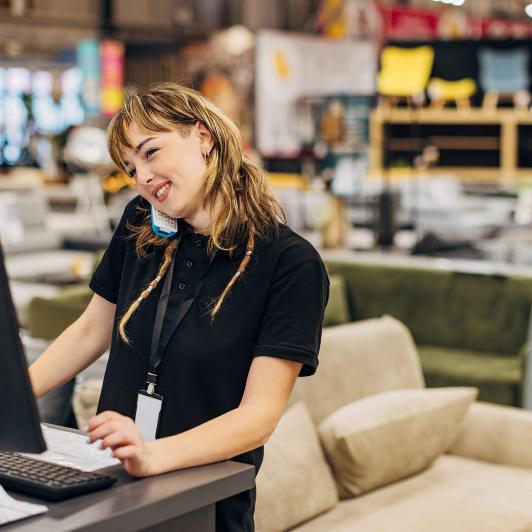 A smiling saleswoman works on a computer in a department store, answers a landline and talks to a customer