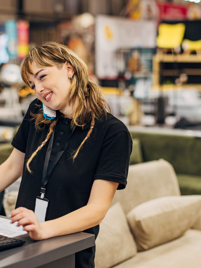 A smiling saleswoman works on a computer in a department store, answers a landline and talks to a customer