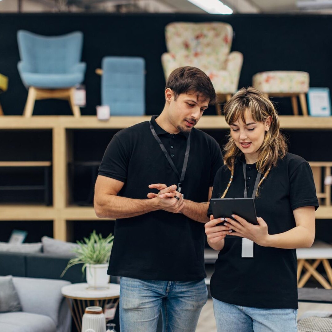 Two salespeople in matching outfits use a digital tablet while working in a furniture store. Teamwork